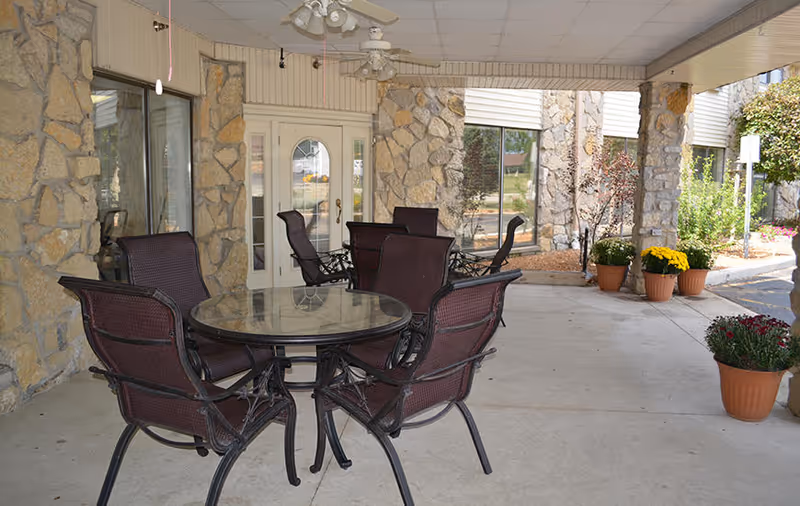 Covered outdoor patio area with a round glass-top table and four metal chairs with woven seats and backs. The patio is supported by stone pillars and has ceiling fans. There are potted plants with flowers placed along the edge of the patio near the building entrance with glass doors and windows.