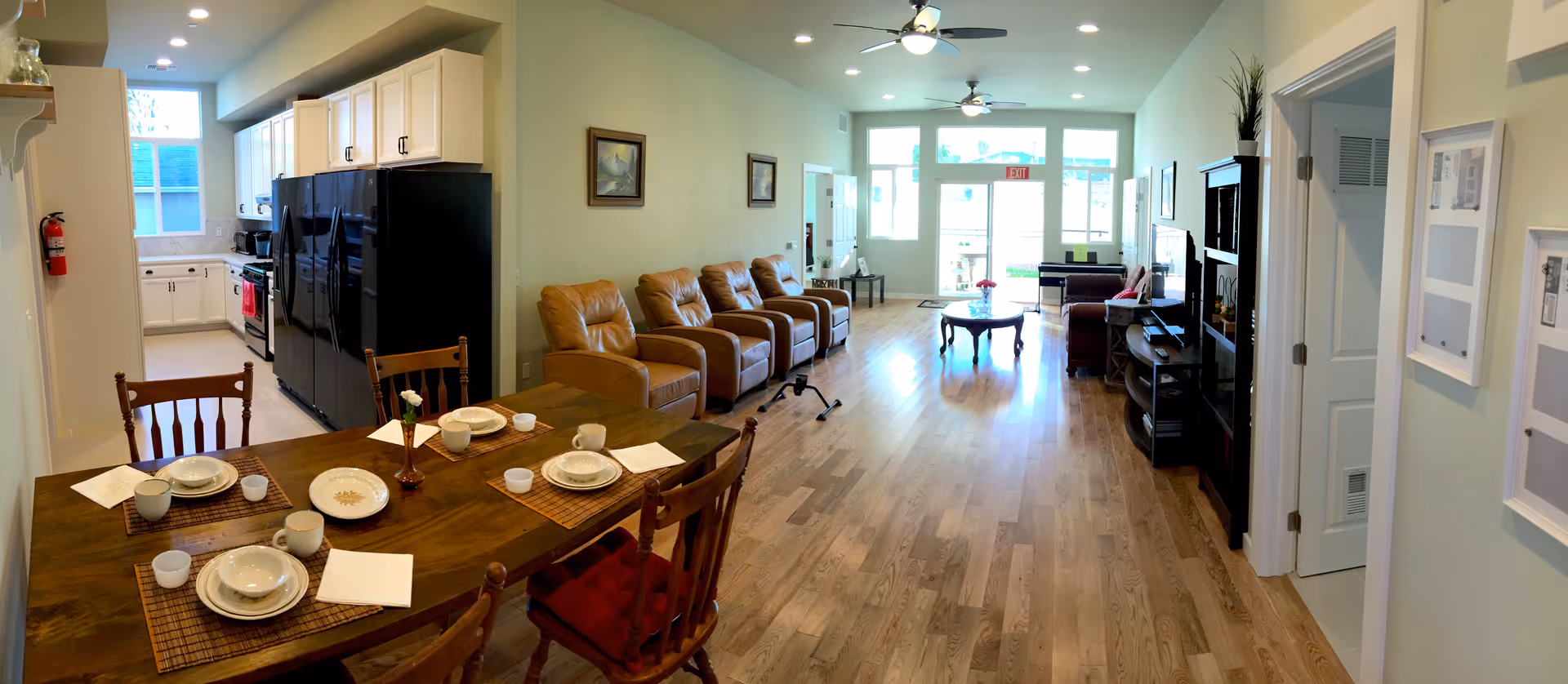 A spacious senior living facility interior featuring a dining area with a wooden table set for four, a kitchen with white cabinets and black appliances, and a living room with four tan recliner chairs, a coffee table, and a TV on a stand. The room has wooden flooring, light-colored walls, and large windows letting in natural light.