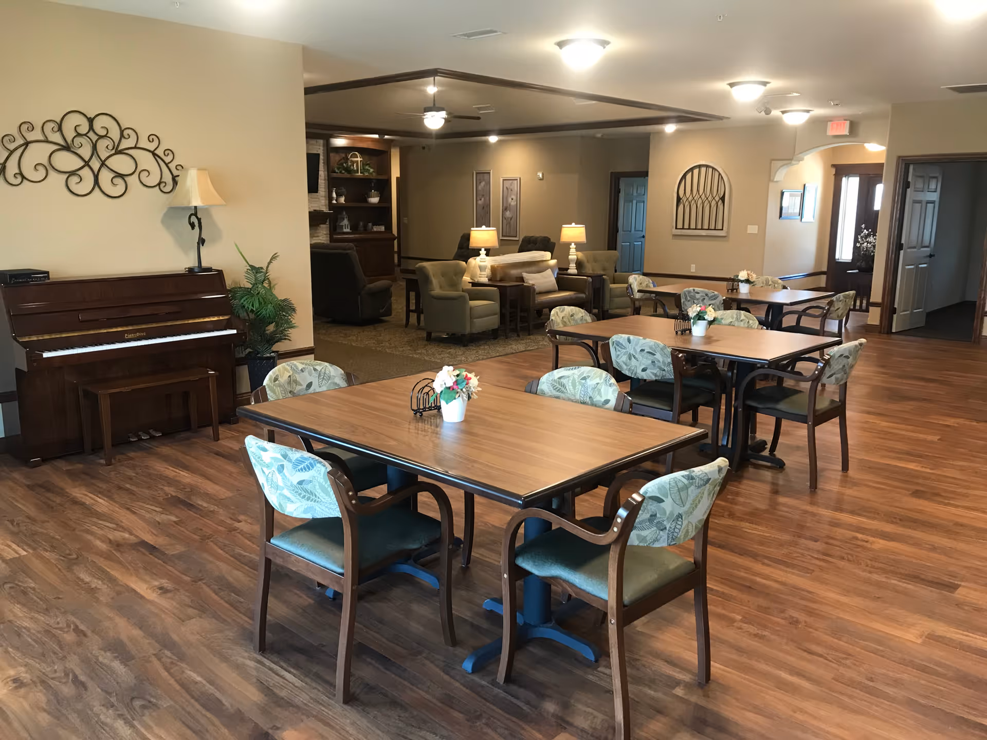 A spacious room with wooden flooring featuring several square tables each surrounded by four chairs with patterned upholstery. In the background, there is a seating area with armchairs and lamps, a piano against the wall, and decorative wall art. The room is well-lit with ceiling lights and has a warm, inviting atmosphere.