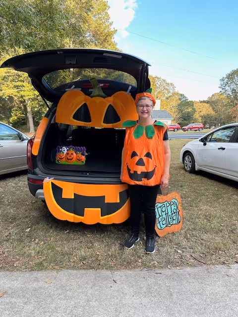 A person dressed in a pumpkin costume stands next to an open car trunk decorated with large pumpkin cutouts. The person is holding a pumpkin-shaped sign that says 'Happy Halloween'. The scene is outdoors in a grassy area with trees and parked cars in the background.