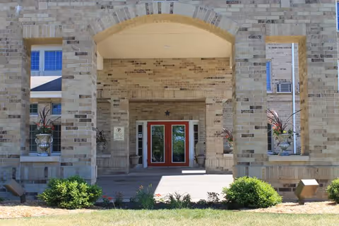 Entrance of a building with a large brick archway and double red doors. There are two large planters with plants on either side of the entrance and small bushes and grass in the foreground.