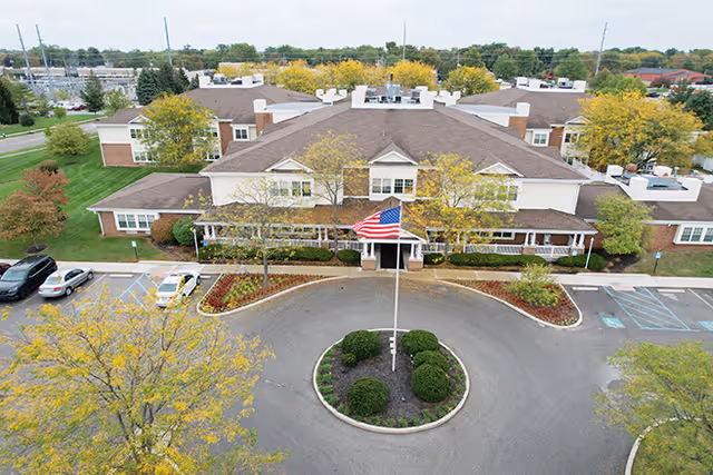Aerial view of Cedar Hill Senior Living facility showing a large two-story building with a brown roof and beige exterior. In front of the building is a circular driveway with a flagpole displaying the American flag, surrounded by neatly trimmed bushes and landscaped flower beds. Several cars are parked in the parking lot adjacent to the building, and trees with autumn foliage are visible around the property.