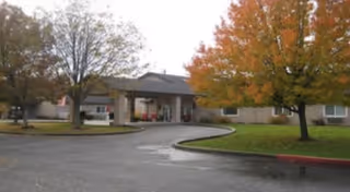 Front entrance of a senior living facility with a covered porte-cochere, circular driveway, and trees showing autumn foliage.