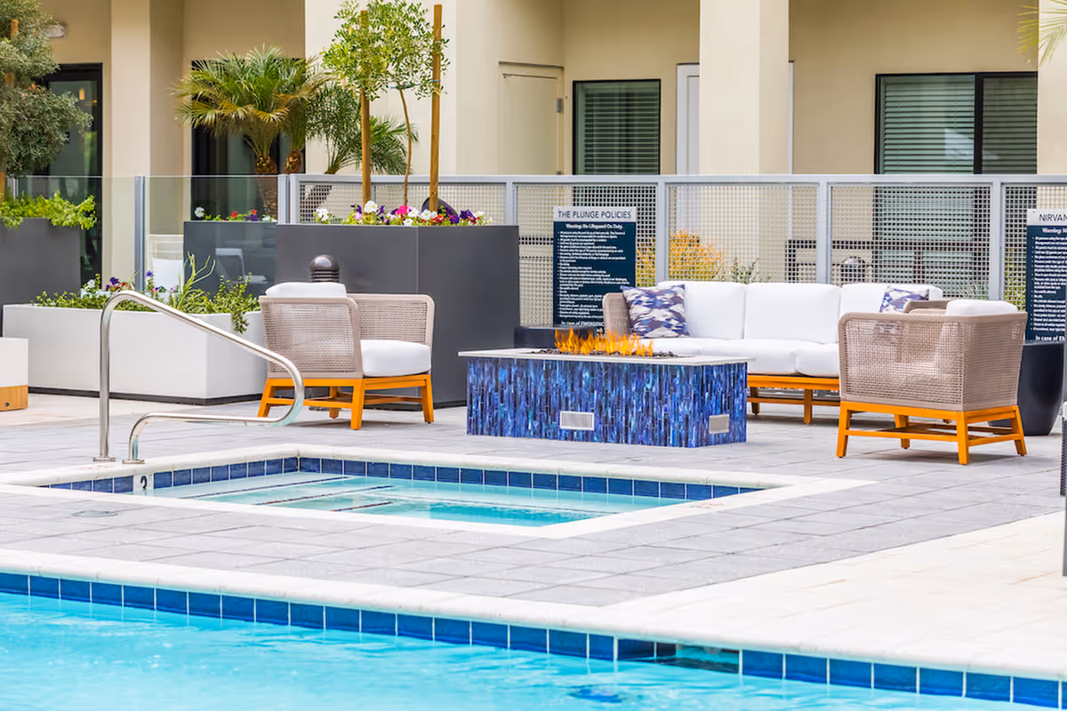 Poolside patio area with a small pool/hot tub, modern lounge chairs, a gas fire pit table and potted plants in front of a building.