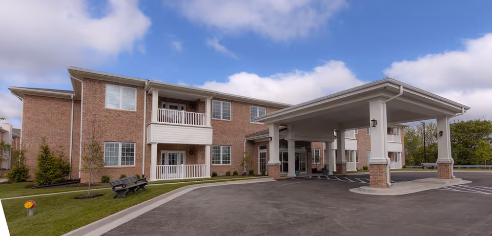 Exterior view of a two-story brick building with white trim and balconies, featuring a covered entrance with large white columns. The building is surrounded by a paved driveway, green grass, small trees, and benches under a partly cloudy sky.