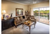 A cozy living room with a brown sofa, an armchair, a wooden coffee table with decorative items, a sideboard with a lamp and framed photos, a painting on the wall, and a sliding glass door leading to a balcony overlooking greenery.