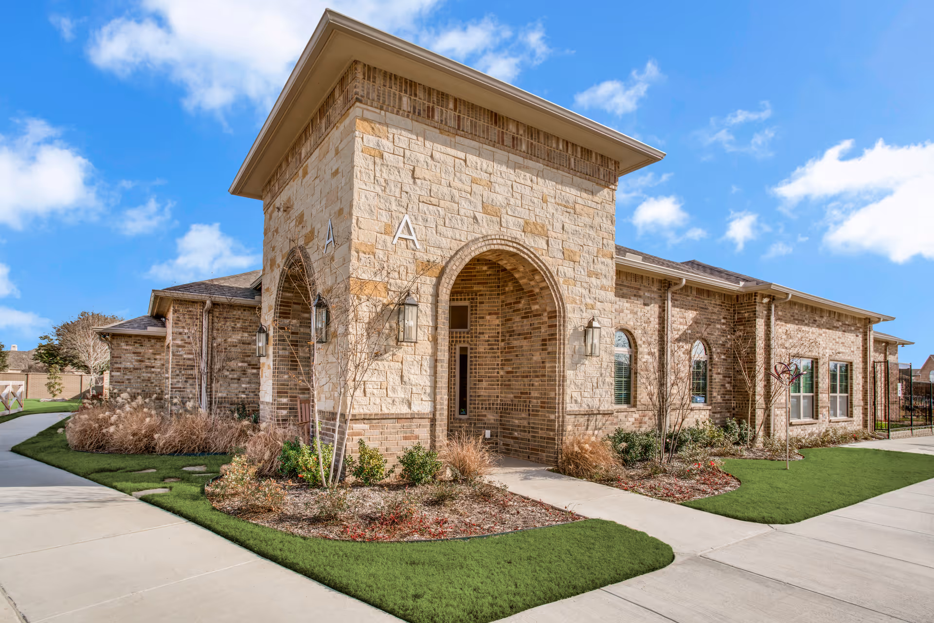 Exterior view of a brick and stone building with an arched entrance, surrounded by landscaped greenery and a clear blue sky with some clouds.