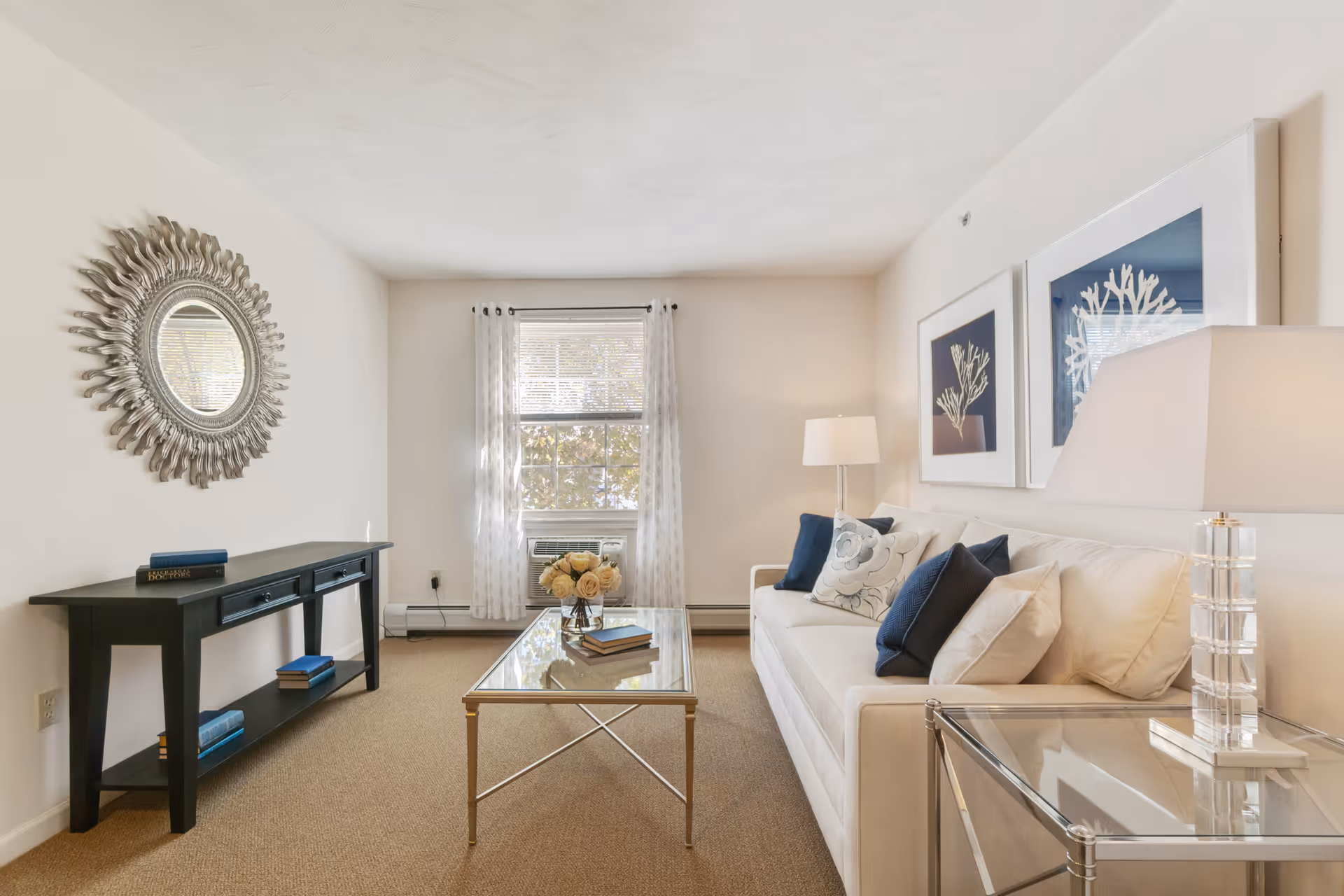 A bright living room with a white sofa adorned with blue and white pillows, a glass coffee table with books and a flower arrangement, a black console table with books, a decorative round mirror on the wall, two framed coral prints above the sofa, a floor lamp, and a window with sheer white curtains.