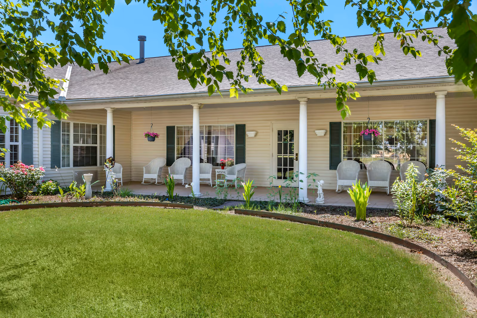 A covered porch area of a building with white columns, white wicker chairs, hanging flower baskets, and a well-maintained garden with green grass and various plants in front. The building has beige siding and green shutters on the windows.