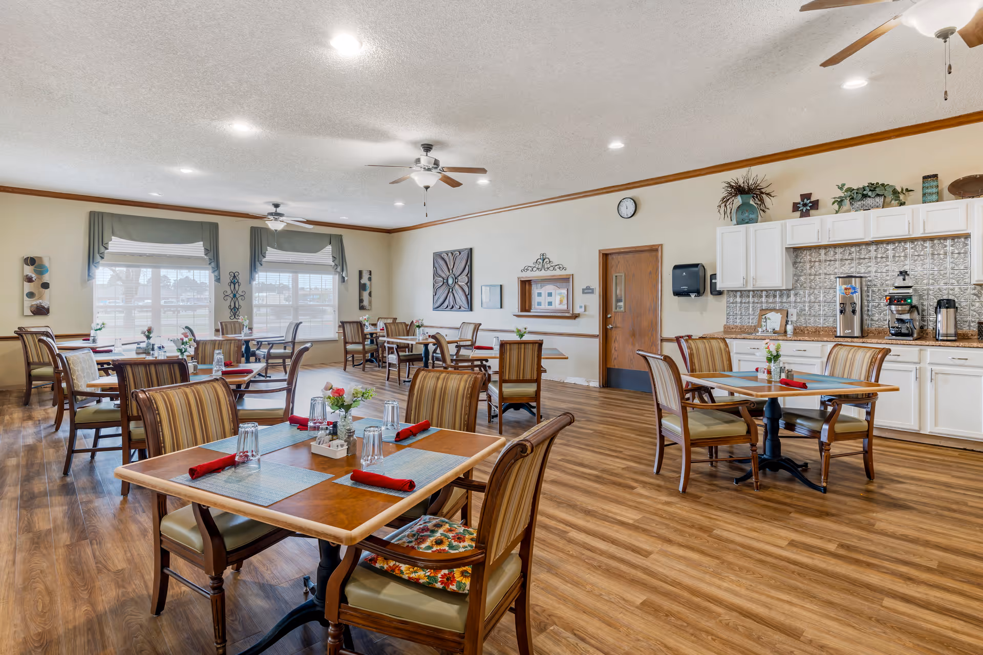 A bright and spacious dining room with several wooden tables and chairs arranged neatly. Each table is set with placemats, glasses, and red napkins. The room features large windows with green valances, ceiling fans, and a wooden floor. There is a counter area with white cabinets, a coffee machine, and decorative plants on top.
