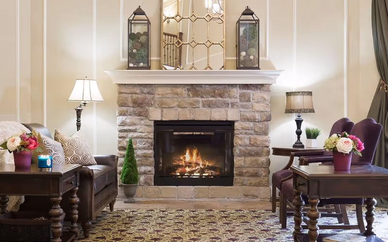 A cozy living room area featuring a stone fireplace with a fire burning inside. On the mantel above the fireplace are two decorative lanterns and a large mirror with a geometric pattern. On either side of the fireplace are side tables with lamps, flower arrangements, and a small potted plant. The room has a patterned carpet and comfortable seating including a leather sofa and upholstered chairs.