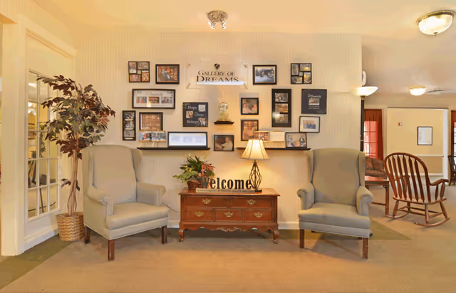 A cozy seating area in a senior living facility with two gray armchairs on either side of a wooden table that has a lamp, a plant, and a 'welcome' sign. Behind the table is a wall decorated with framed photos and inspirational quotes under a sign that reads 'Gallery of Dreams.' A potted plant is placed to the left of the chairs, and a rocking chair is visible in the background.
