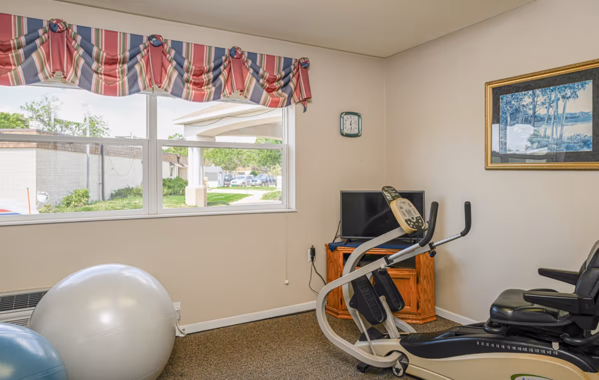 A small exercise room with a large window covered by a striped valance, an exercise bike, two large exercise balls, a small TV on a wooden stand, a wall clock, and a framed landscape painting on the wall.