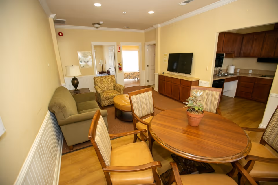 Interior view of a senior living facility common area featuring a round wooden table with four chairs, a green sofa, a patterned armchair, a round ottoman, a wall-mounted flat-screen TV above a wooden cabinet, and a kitchen area with wooden cabinets and appliances visible in the background.