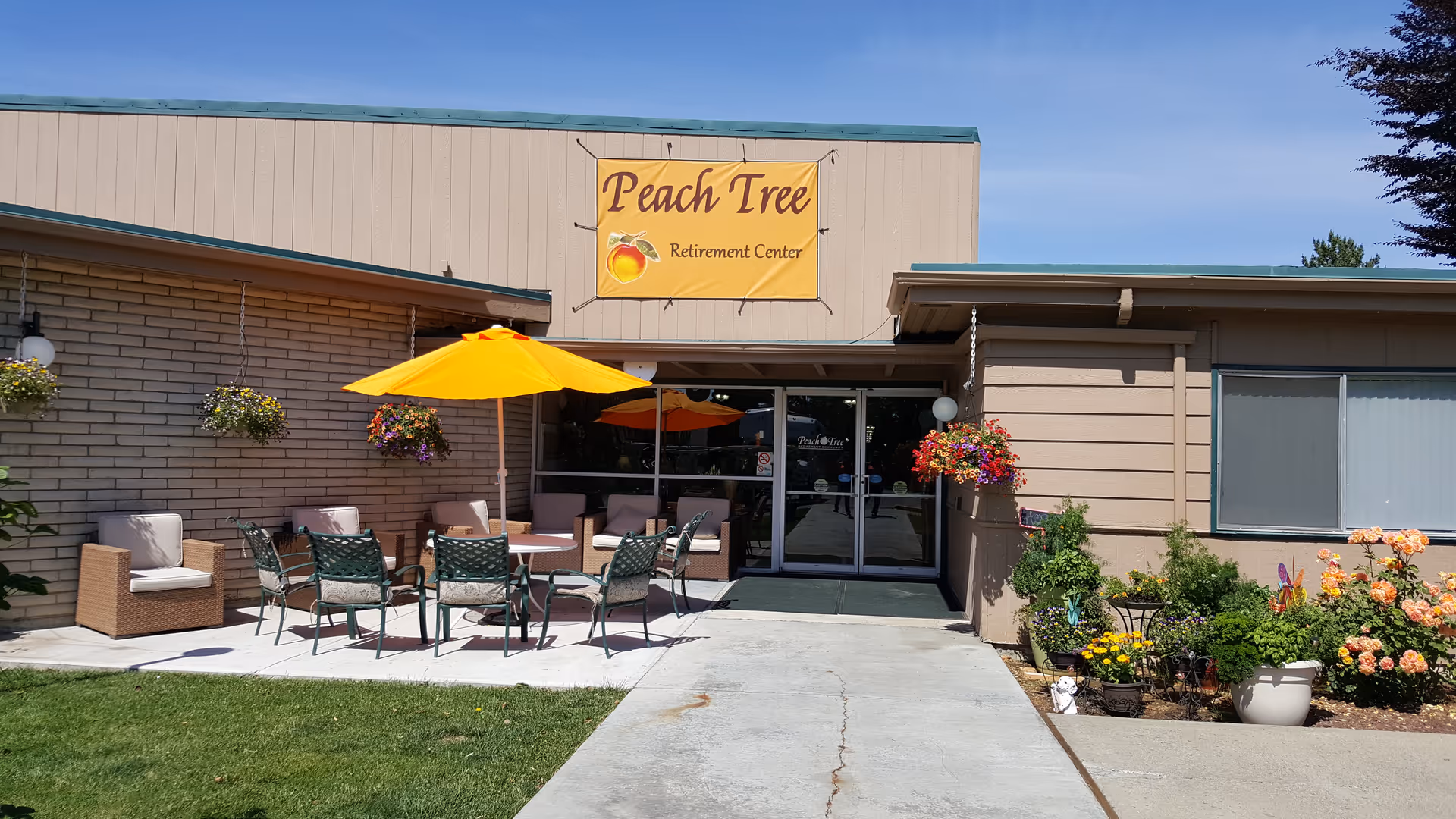 Outdoor patio area at Peach Tree Retirement Center with several chairs and tables under bright yellow umbrellas, surrounded by hanging flower baskets and garden plants near the entrance of the building.