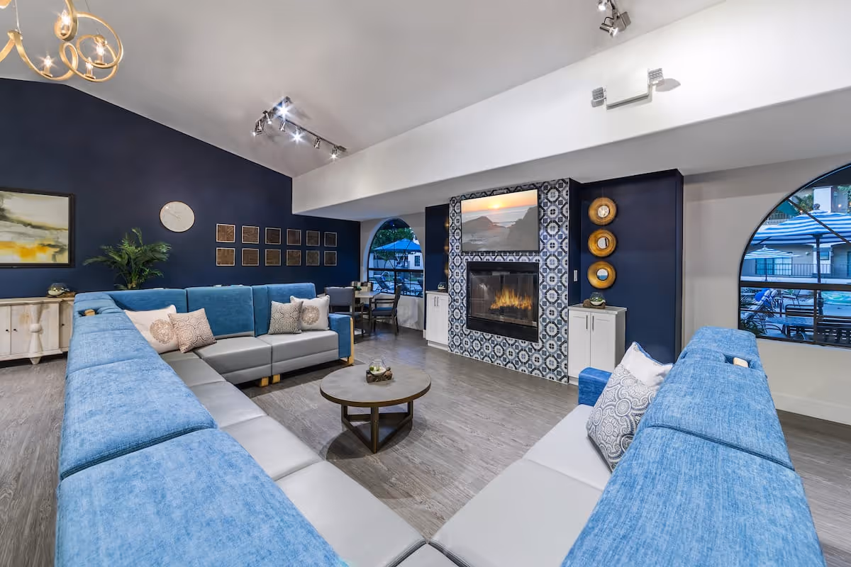 A modern living room with a large U-shaped blue and gray sectional sofa surrounding a round coffee table. The room features a fireplace with a decorative tiled surround and a TV mounted above it. There are two arched windows showing an outdoor area with umbrellas and seating. The walls are painted dark blue and white, with various wall decorations and a plant on a sideboard.