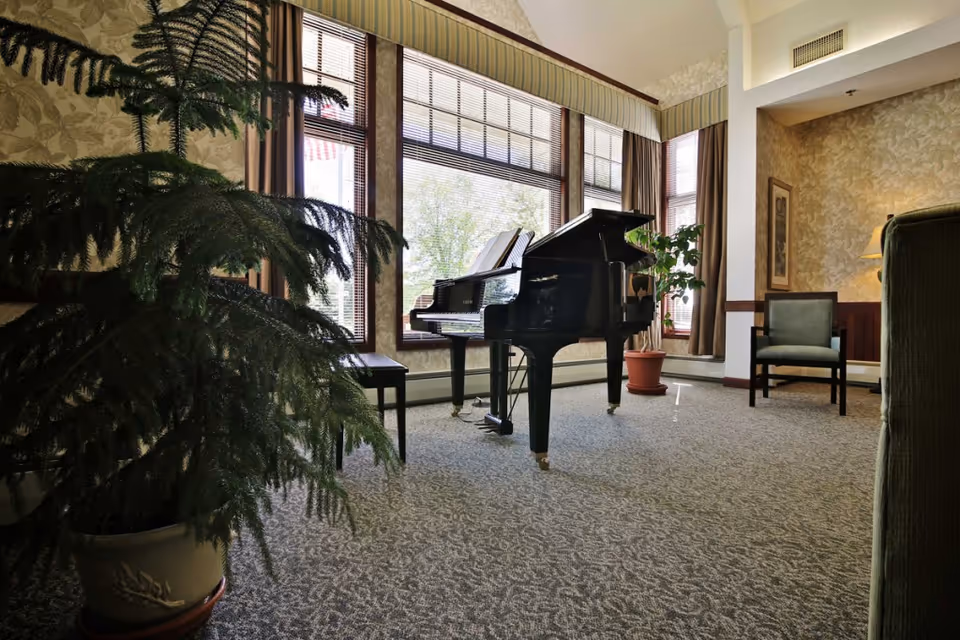 A cozy living room area in Oak Meadows Senior Living featuring a black grand piano with sheet music on the stand, a large potted plant in the foreground, another potted plant near the windows, a green upholstered chair, and warm patterned wallpaper. Large windows with blinds and curtains allow natural light to fill the room.