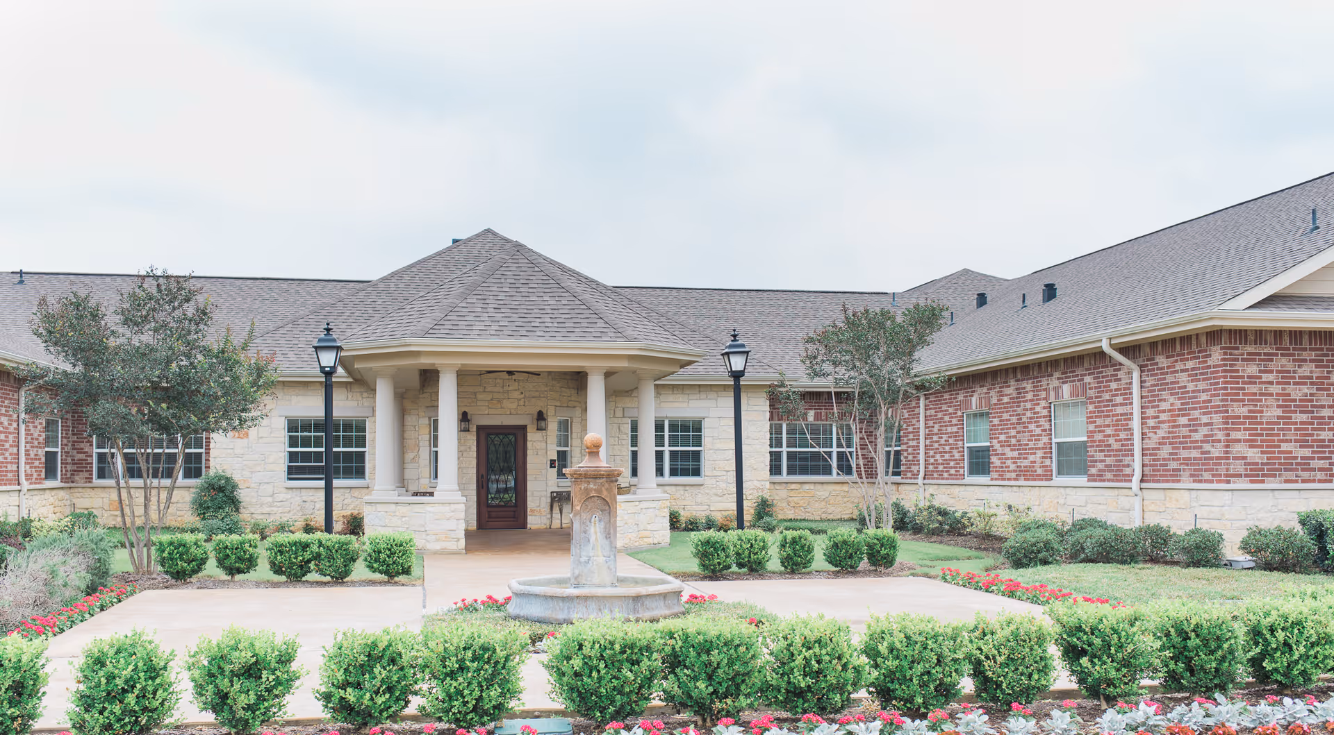 Front exterior view of The Heritage at Eldridge Parkway building with a covered entrance supported by columns, a central fountain, manicured bushes, flower beds, and two black lamp posts on either side of the walkway.