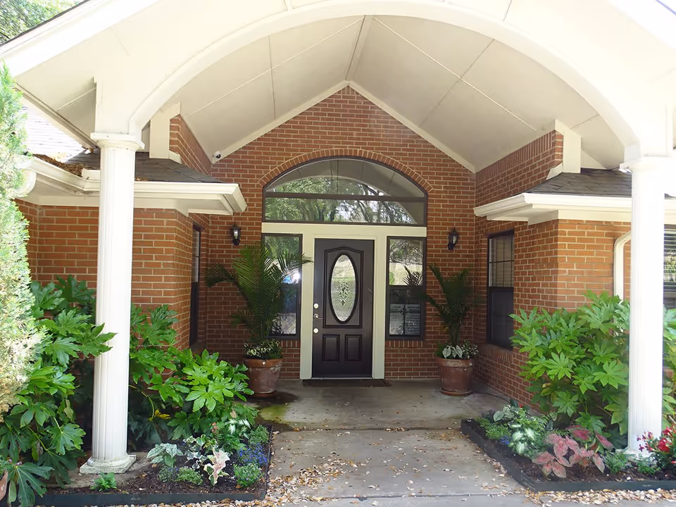 Entrance to a brick building with a covered porch supported by white columns. The door is dark with an oval glass panel, flanked by two windows and potted plants on either side. There are green shrubs and plants along the walkway leading to the door.