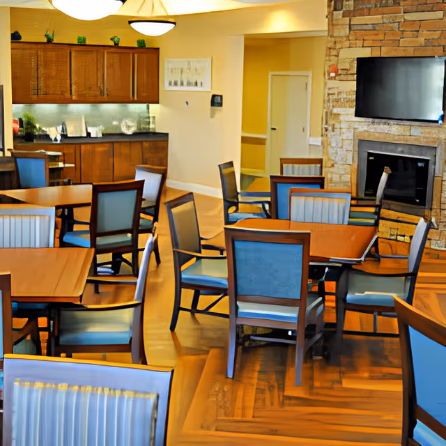 A dining room with multiple wooden tables and chairs with blue cushions. The room features a stone fireplace with a mounted flat-screen TV above it. There are wooden cabinets along one wall and a doorway leading to another area. The floor is wooden and the room is well-lit with ceiling lights.