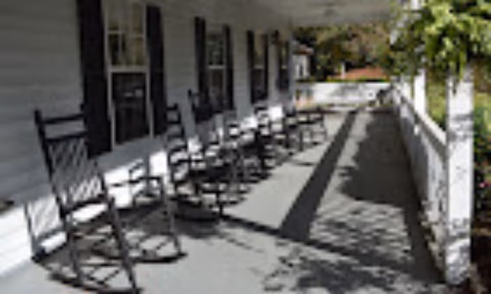 A long porch with several black rocking chairs lined up against the white exterior wall of a building, with windows and black shutters. The porch has a white railing and overlooks a garden area with greenery.