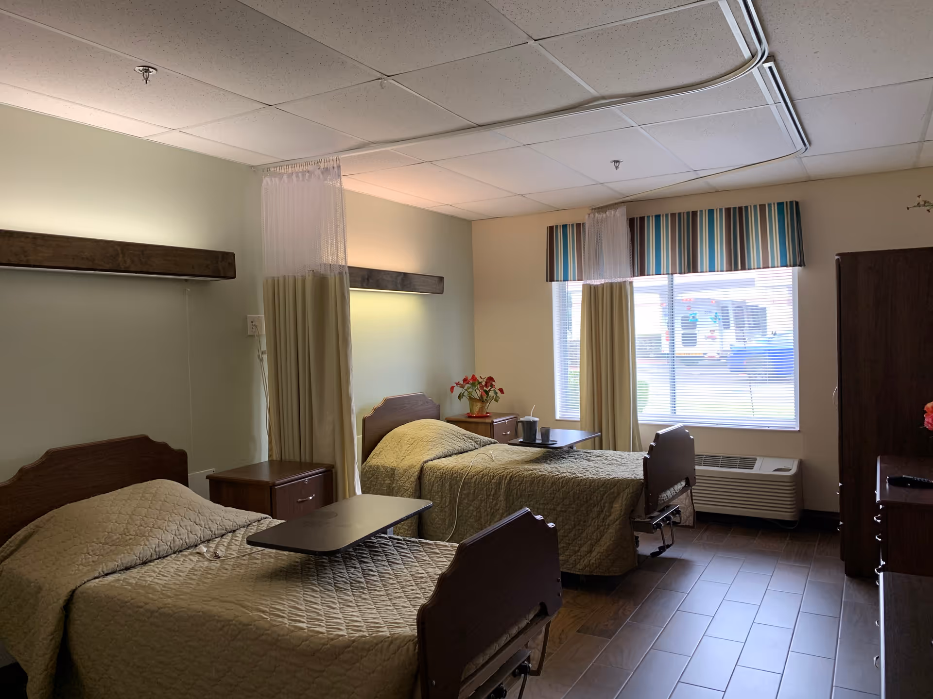 A nursing home room with two hospital-style beds covered in beige quilts, each with an overbed table. There are privacy curtains hanging from ceiling tracks between the beds and near the window. A window with striped valance lets in natural light. The room has wooden furniture including nightstands and a dresser, and a small plant on one nightstand.