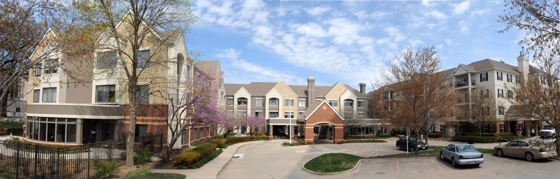 Exterior view of a multi-story retirement facility building with a covered entrance, surrounded by trees with some spring blossoms and a parking area with several cars parked. The sky is partly cloudy.