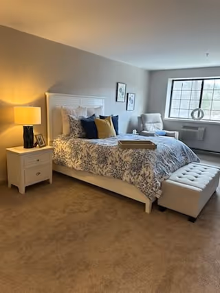 A cozy bedroom with a white bed frame and floral bedding, a nightstand with a lamp and picture frame, a cushioned bench at the foot of the bed, a comfortable armchair near a window, and two framed artworks on the wall.