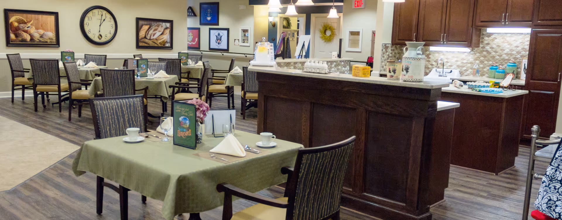 A dining area in a senior living facility with several tables covered in green tablecloths, each set with cups, glasses, napkins, and menus. The room features wooden flooring, a large clock on the wall, framed pictures, and a kitchen area with dark wood cabinets and a countertop. The space is well-lit with ceiling lights and has a cozy, welcoming atmosphere.