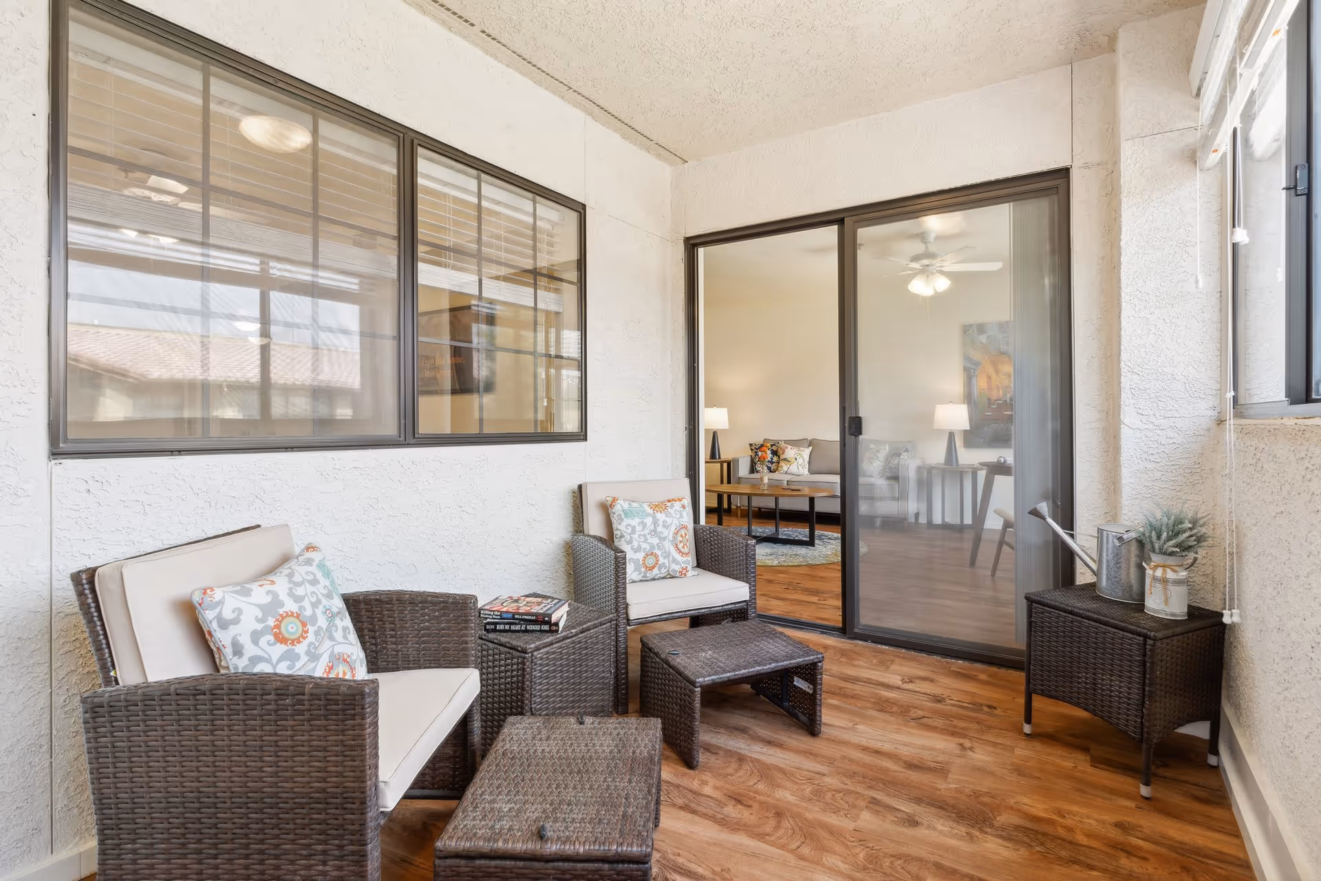 A cozy enclosed patio area with two cushioned wicker chairs, a small wicker table with books, and a wicker ottoman. The patio has wood flooring and white textured walls. A sliding glass door leads to a living room with a sofa, lamps, and a ceiling fan. A watering can and a small plant are placed on a wicker side table near a window.