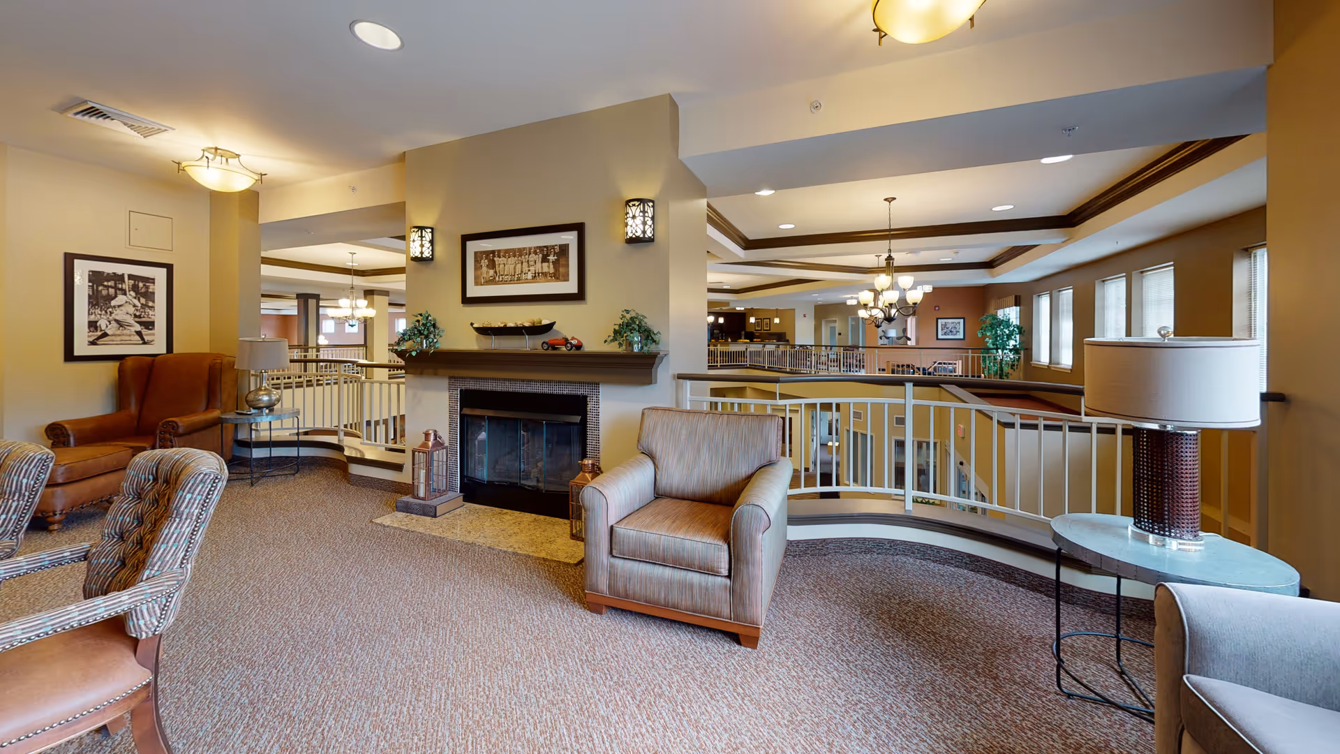 A cozy senior living facility common area with comfortable armchairs arranged around a fireplace. The room features warm beige walls, framed artwork, a carpeted floor, and a railing overlooking a lower level. There are ceiling lights and a table lamp providing soft lighting.