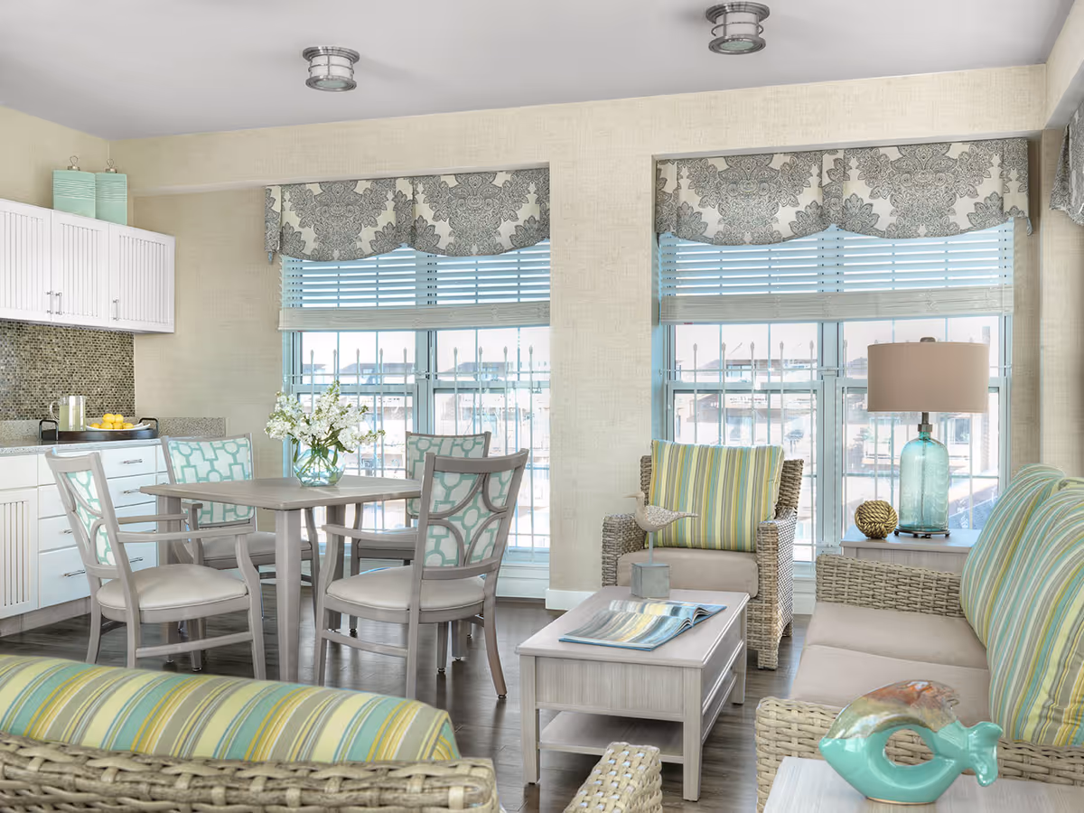 A bright and airy senior living common area featuring a small dining table with four chairs, a cozy seating area with wicker furniture and striped cushions, decorative items including a glass vase with white flowers, a table lamp with a blue glass base, and patterned window valances over large windows with blinds.