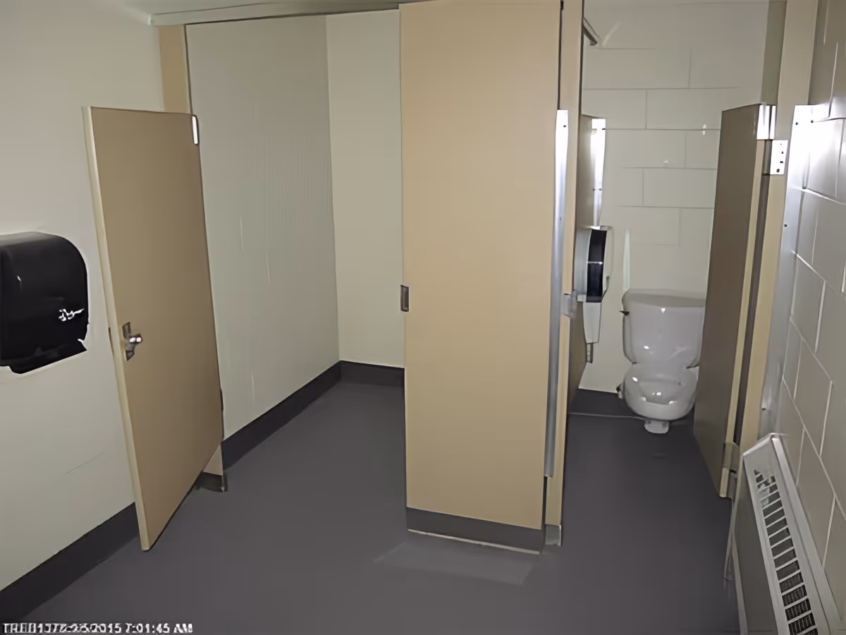 Interior view of a public restroom with beige stall doors, a white toilet inside one stall, a black paper towel dispenser mounted on the wall, and a wall-mounted soap dispenser next to the toilet.