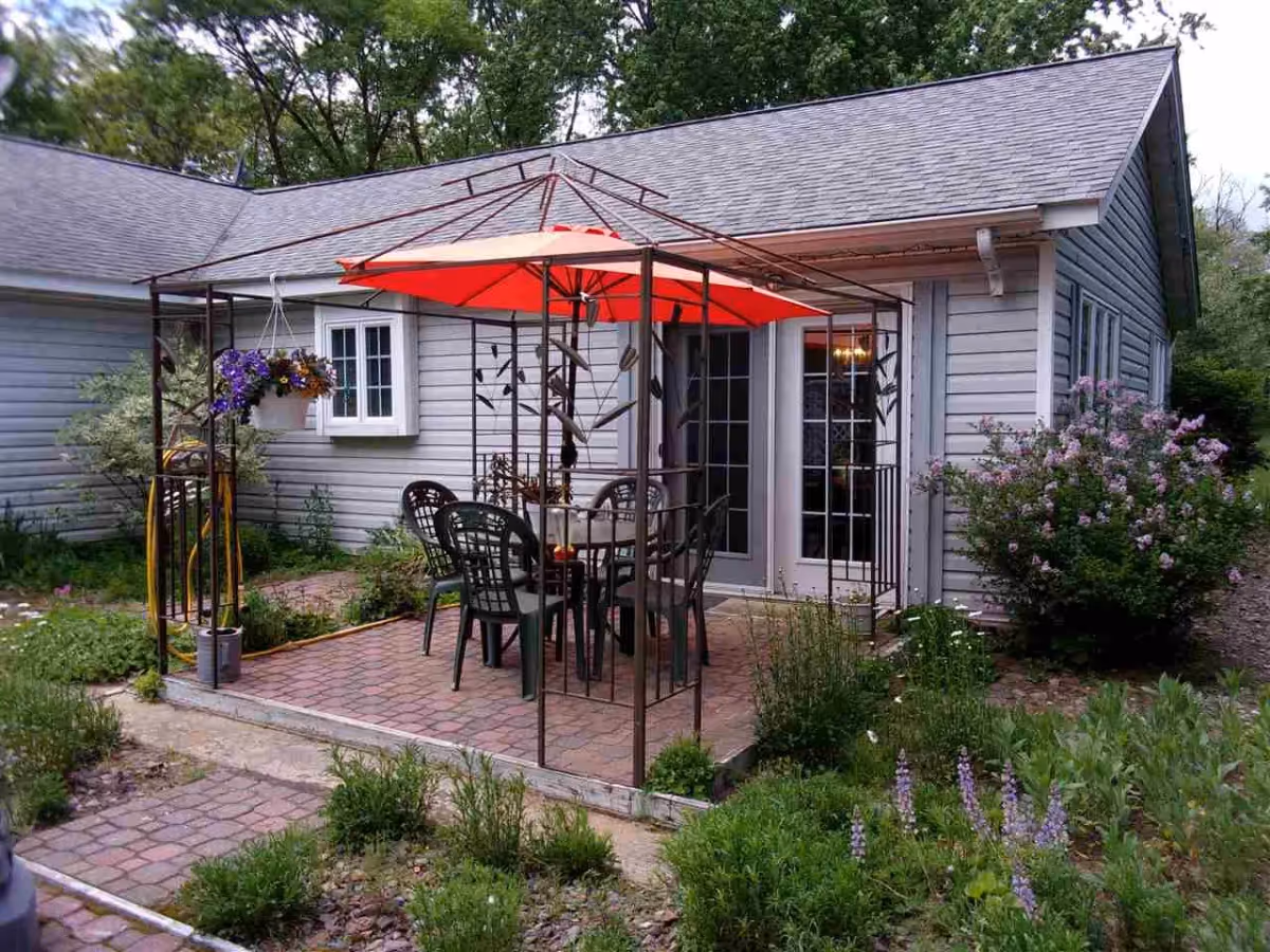Outdoor patio area with a metal gazebo frame and a red umbrella over a table surrounded by six black plastic chairs. The patio is paved with bricks and is adjacent to a gray building with white-framed windows and a glass door. There are various green plants and flowers around the patio.