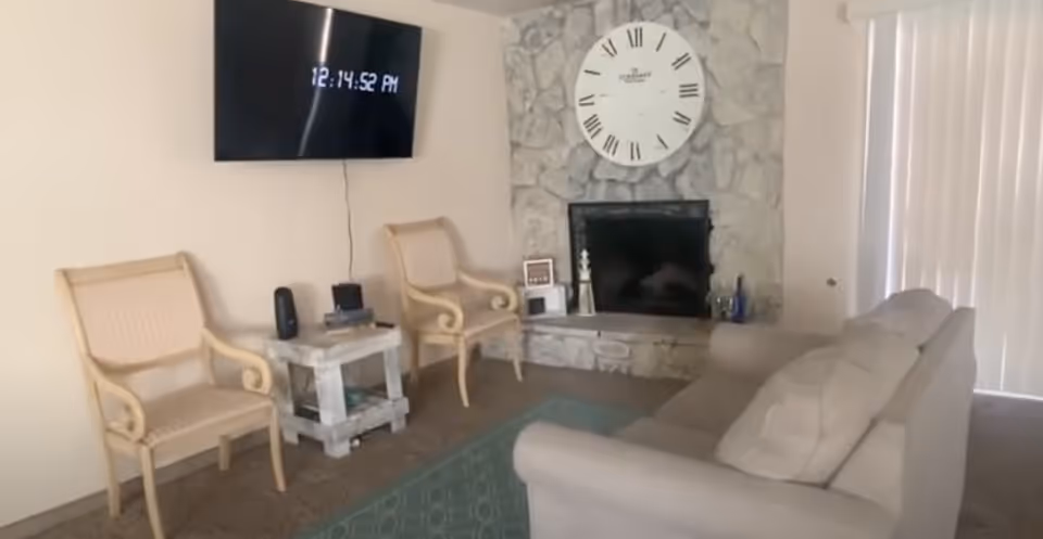 Living room with a sofa and two chairs facing a stone fireplace topped by a large clock and a wall-mounted TV displaying the time.