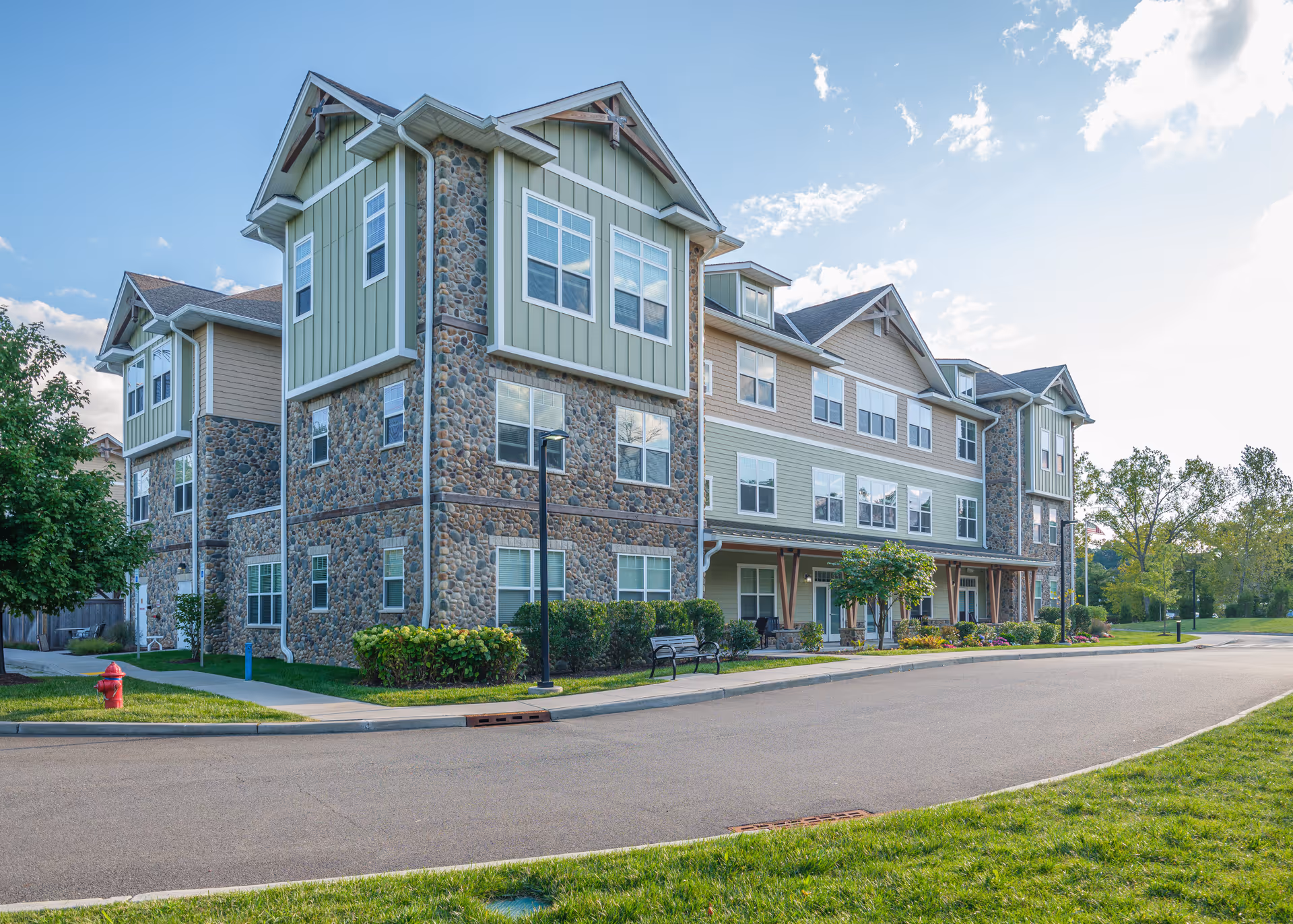 Exterior view of a multi-story senior living facility building with stone and green siding, multiple windows, a sidewalk, a bench, a fire hydrant, and landscaped greenery under a partly cloudy sky.