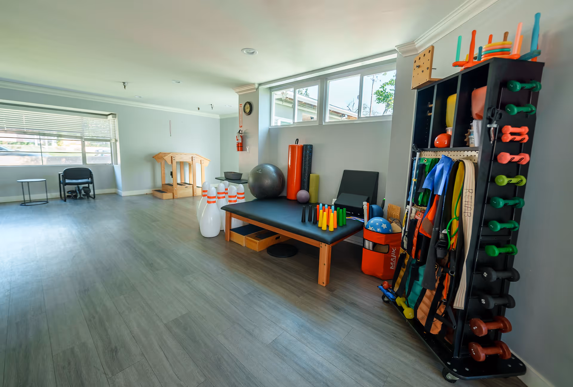 Bright therapy/exercise room with a treatment table, exercise balls, bowling pins and a rack of dumbbells and fitness equipment.