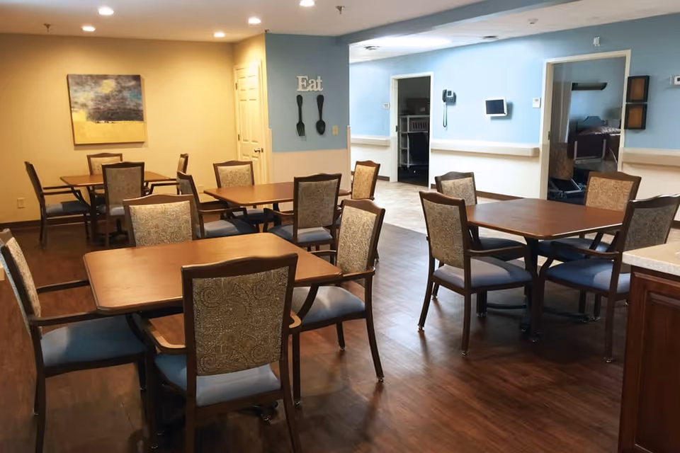 A dining room in a senior living facility with several wooden tables and cushioned chairs arranged neatly on a wooden floor. The walls are painted in light blue and beige tones, with a decorative wall piece featuring the word 'Eat' and large fork and spoon silhouettes. There is a painting on one wall and an open doorway leading to another room.