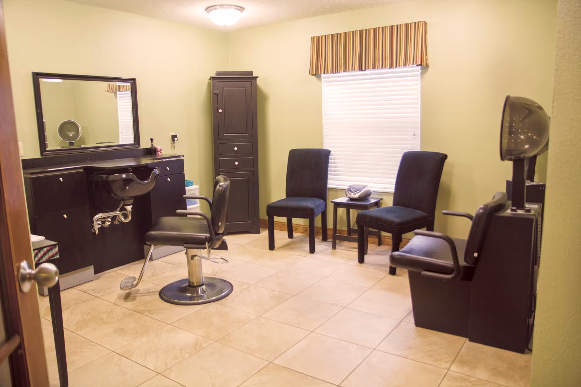 Interior view of a hair salon room with a black salon chair in front of a black sink and counter with a large mirror. There is a tall black cabinet against a light green wall, two black chairs with a small table between them under a window with closed blinds and a striped valance. A hair dryer chair is positioned on the right side of the room.