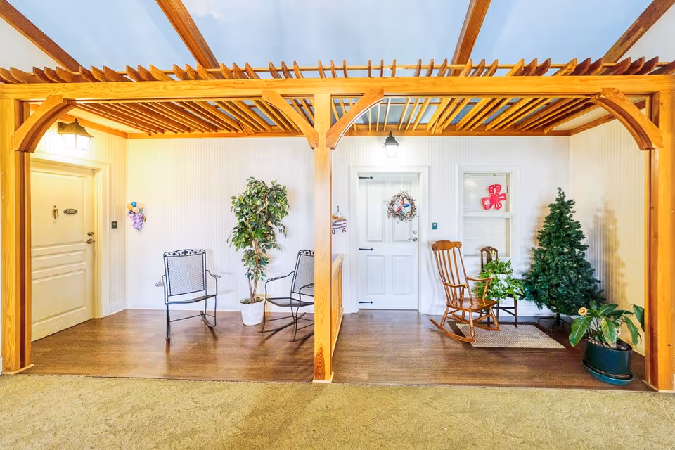 Indoor seating area with wooden pergola-style ceiling beams, two black metal chairs and a potted plant on the left side, and a wooden rocking chair, small wooden chair, potted plants, and an artificial Christmas tree on the right side. There are two white doors with decorative wreaths and a window with a red butterfly decoration on the wall behind the seating. The floor is wooden and the surrounding area has carpet.