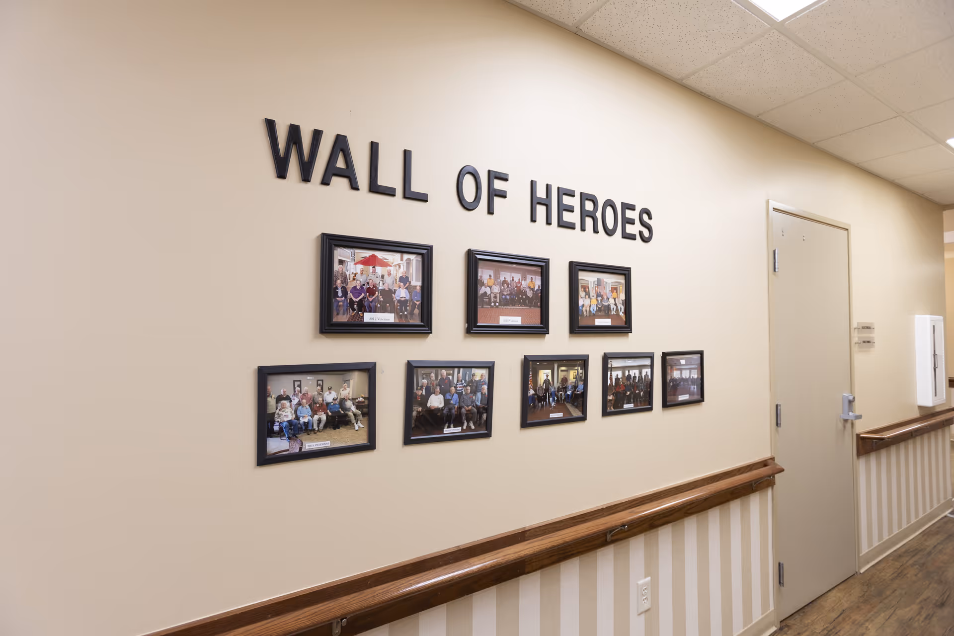 A hallway wall display titled 'Wall of Heroes' featuring eight framed photographs of groups of people, likely residents or staff, in a senior living facility. The wall is beige with a wooden handrail and a closed door is visible at the end of the hallway.