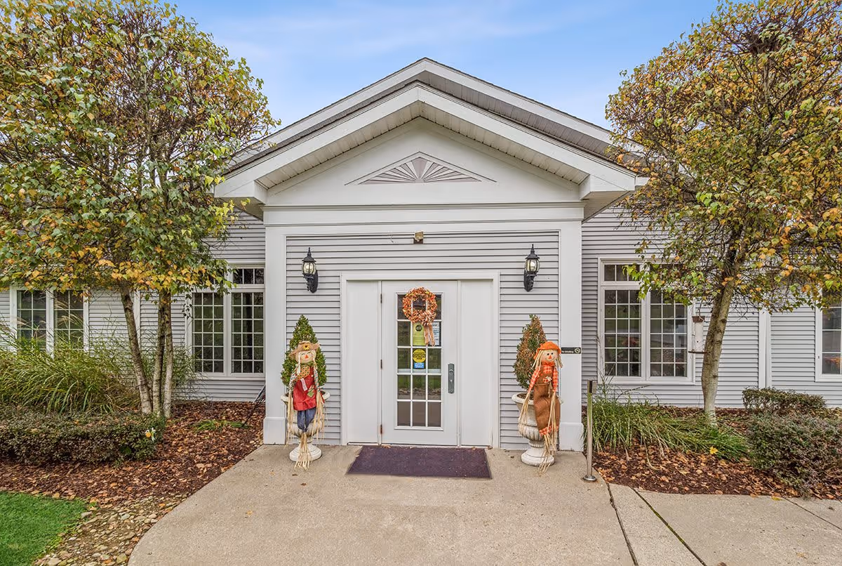 Front entrance of a building with white siding and a peaked roof over the door. Two decorative scarecrow figures stand on either side of the door, which has a fall-themed wreath. There are two small evergreen trees in planters next to the door, and trees with autumn foliage flank the entrance. The walkway leading to the door is concrete.