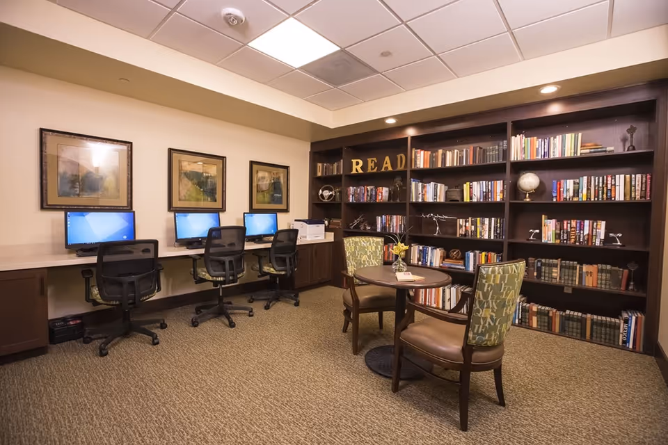 A cozy library and computer room featuring a large dark wood bookshelf filled with books and decorative items. In front of the bookshelf is a small round table with two upholstered chairs. Along one wall, there is a long desk with three computer workstations and office chairs. The room has framed artwork on the walls and recessed lighting in the ceiling.