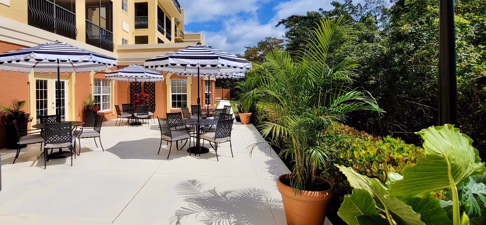 Sunny outdoor patio with round tables, striped umbrellas, metal chairs, and potted palms next to a light-colored building.