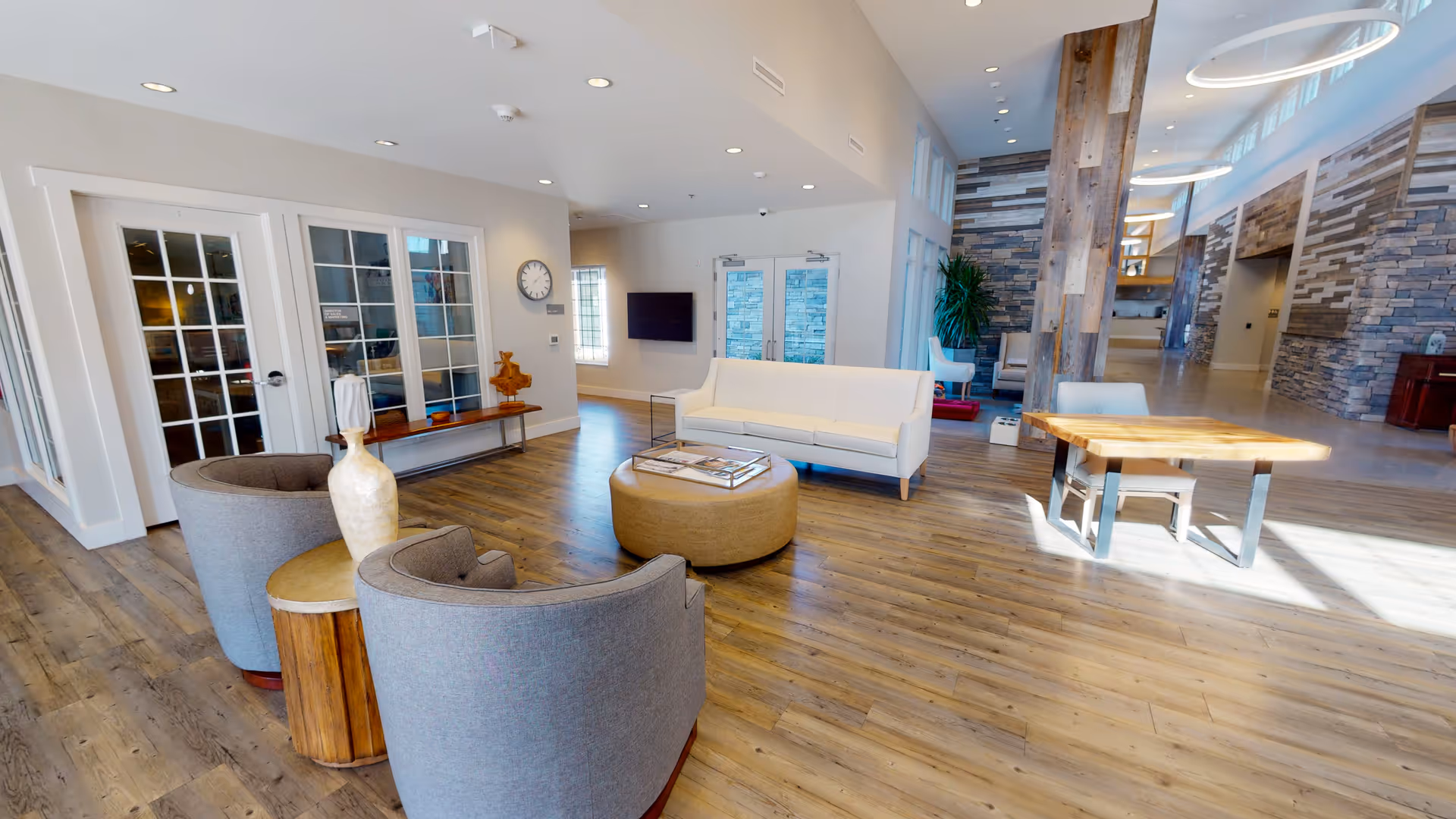 A spacious and modern living room area with wooden flooring, featuring a white sofa, two gray armchairs, a round ottoman, a wooden side table with a decorative vase, and a wooden table with chairs. The room has large windows and glass doors, stone accent walls, and circular ceiling lights.