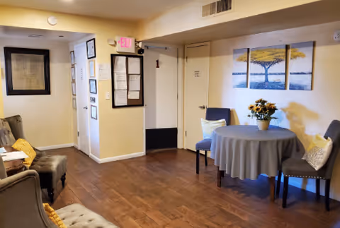 Small assisted-living common room with chairs, a round table with flowers, wall art, and bulletin boards on hardwood floors.