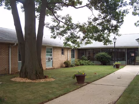 Courtyard with a large tree, grassy lawn, potted plants and a sidewalk leading to the entrance of a single-story building.