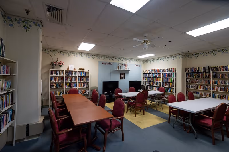 A library or common room with multiple bookshelves filled with books along the walls. There are several tables with red cushioned chairs arranged around them. The room has a ceiling fan and fluorescent lighting. On the far wall, there are two televisions and a sign that reads 'Ingleside Bowling Lanes'.