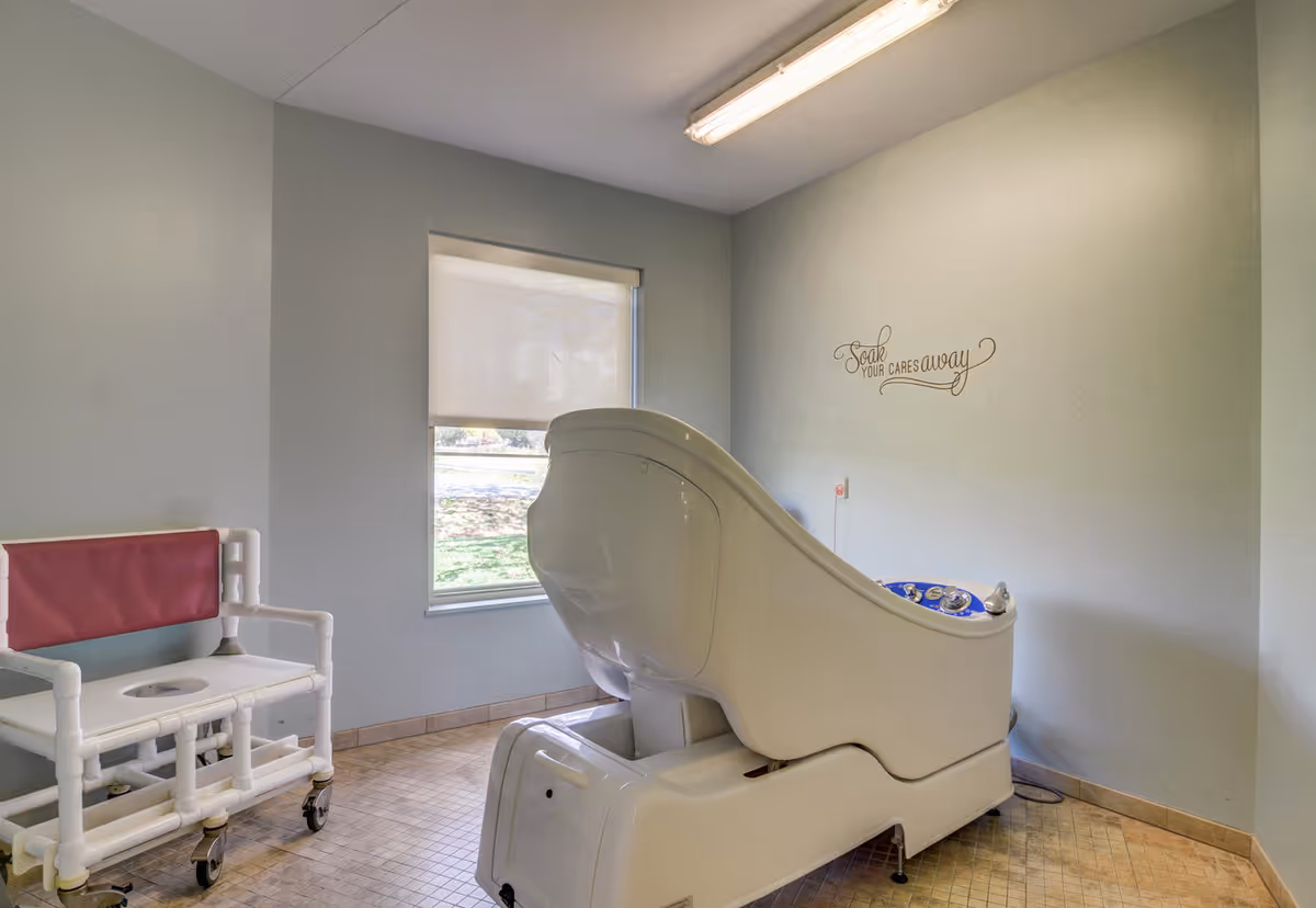 A small room with a large white medical or therapeutic bathtub and a white chair with a red backrest on wheels. There is a window with a roller shade partially down, and a wall decal that reads 'Soak your cares away'. The floor is tiled and the walls are painted light gray.