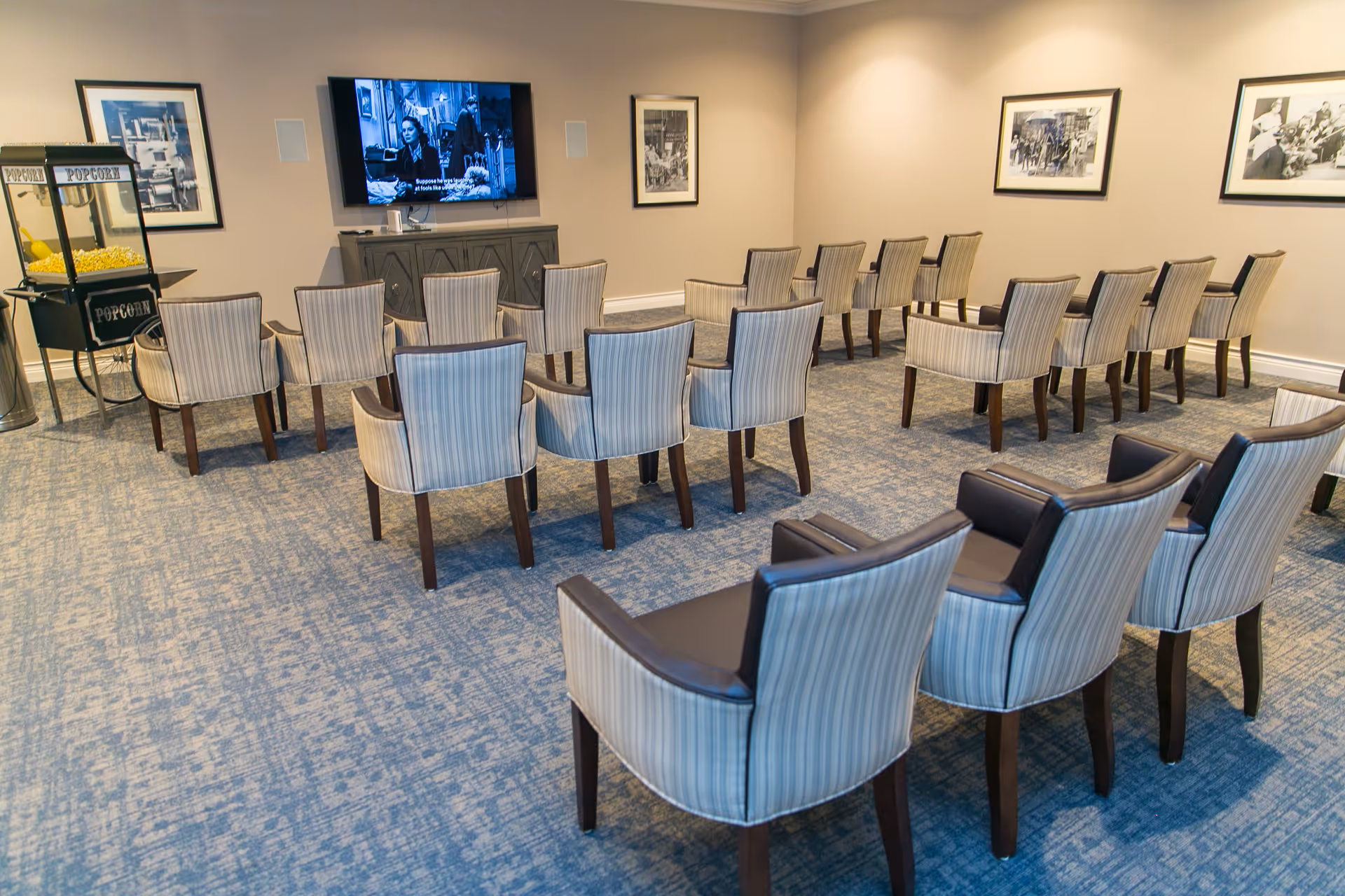 A small theater-style room with multiple rows of striped upholstered chairs facing a wall-mounted flat screen TV. There is a popcorn machine on the left side of the room and black-and-white framed photographs on the beige walls.